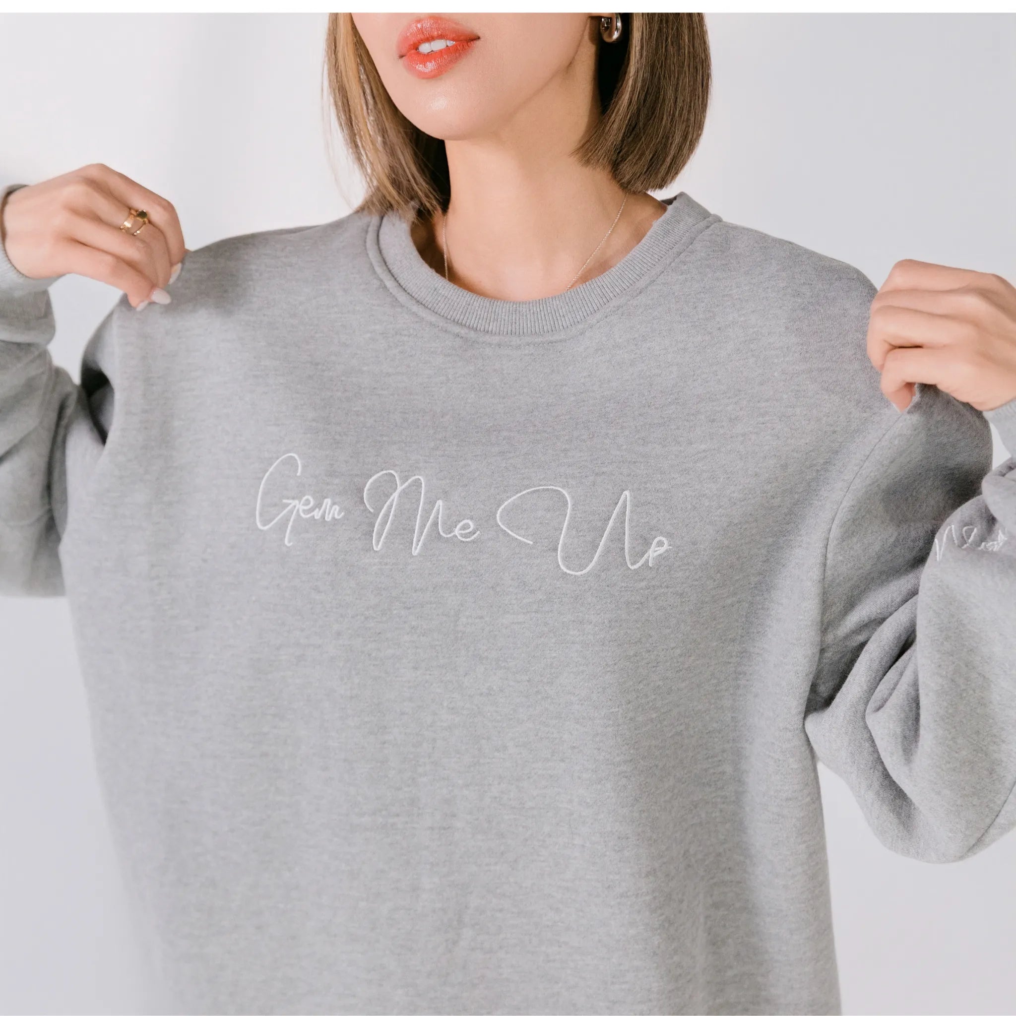 Gray sweatshirt with 'Gen Me Up' text held by a person on a light gray background