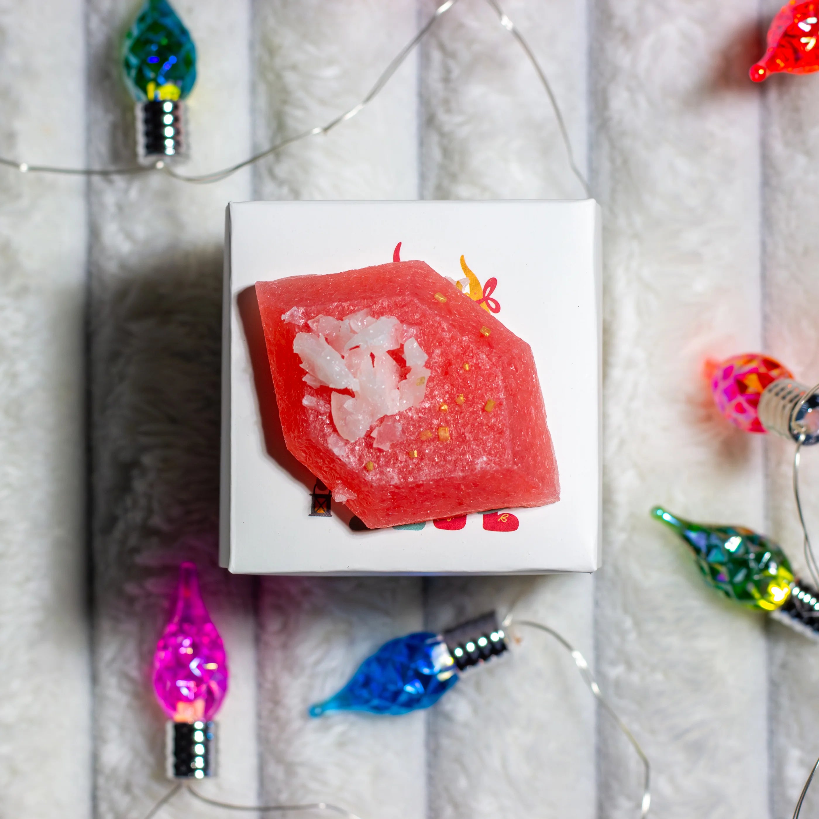 Red soap bar with decorative elements on a white card, surrounded by colorful Christmas lights.