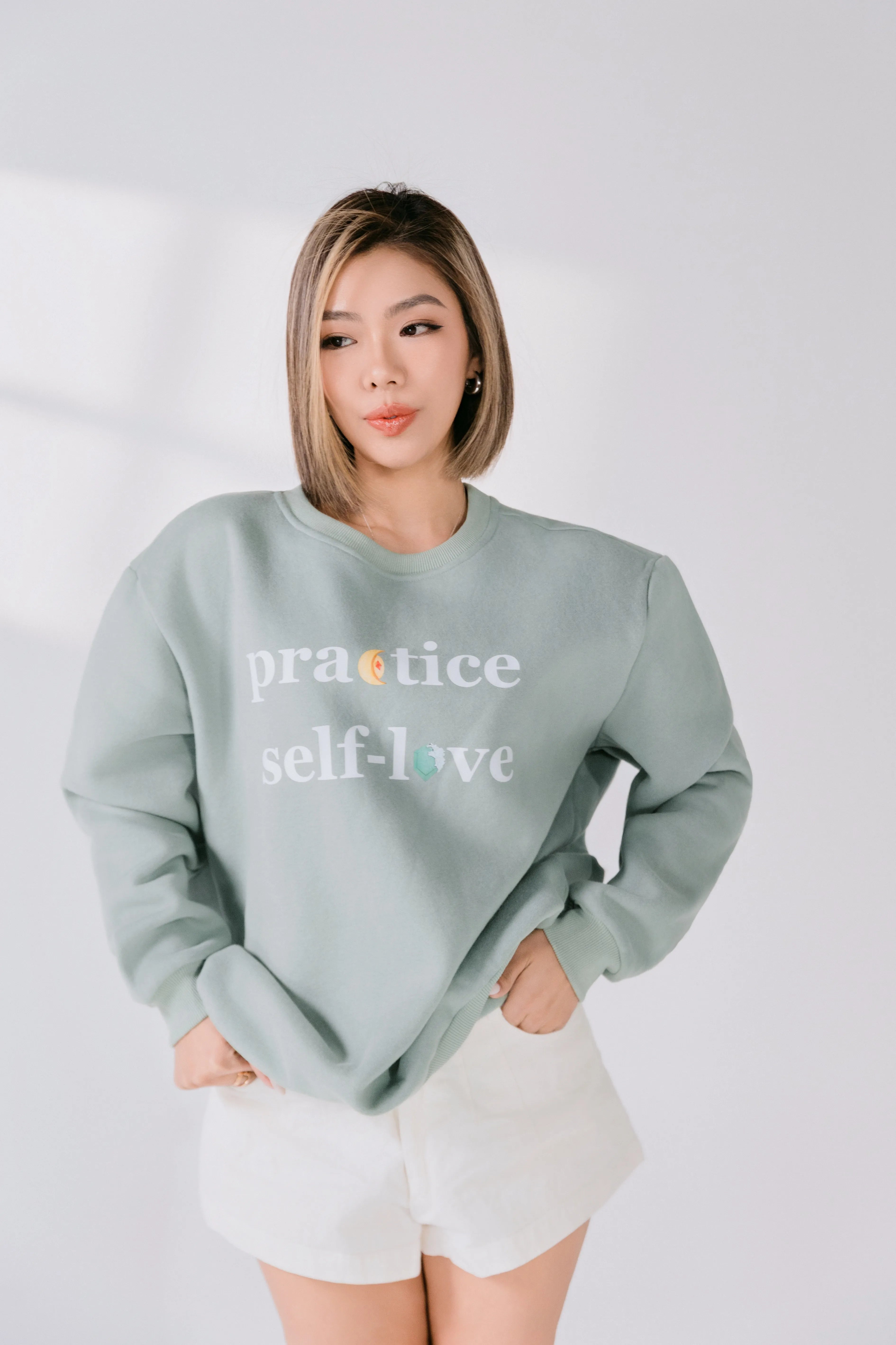 Three-quarter view of sage green Practice Self-Love sweatshirt styled casually.