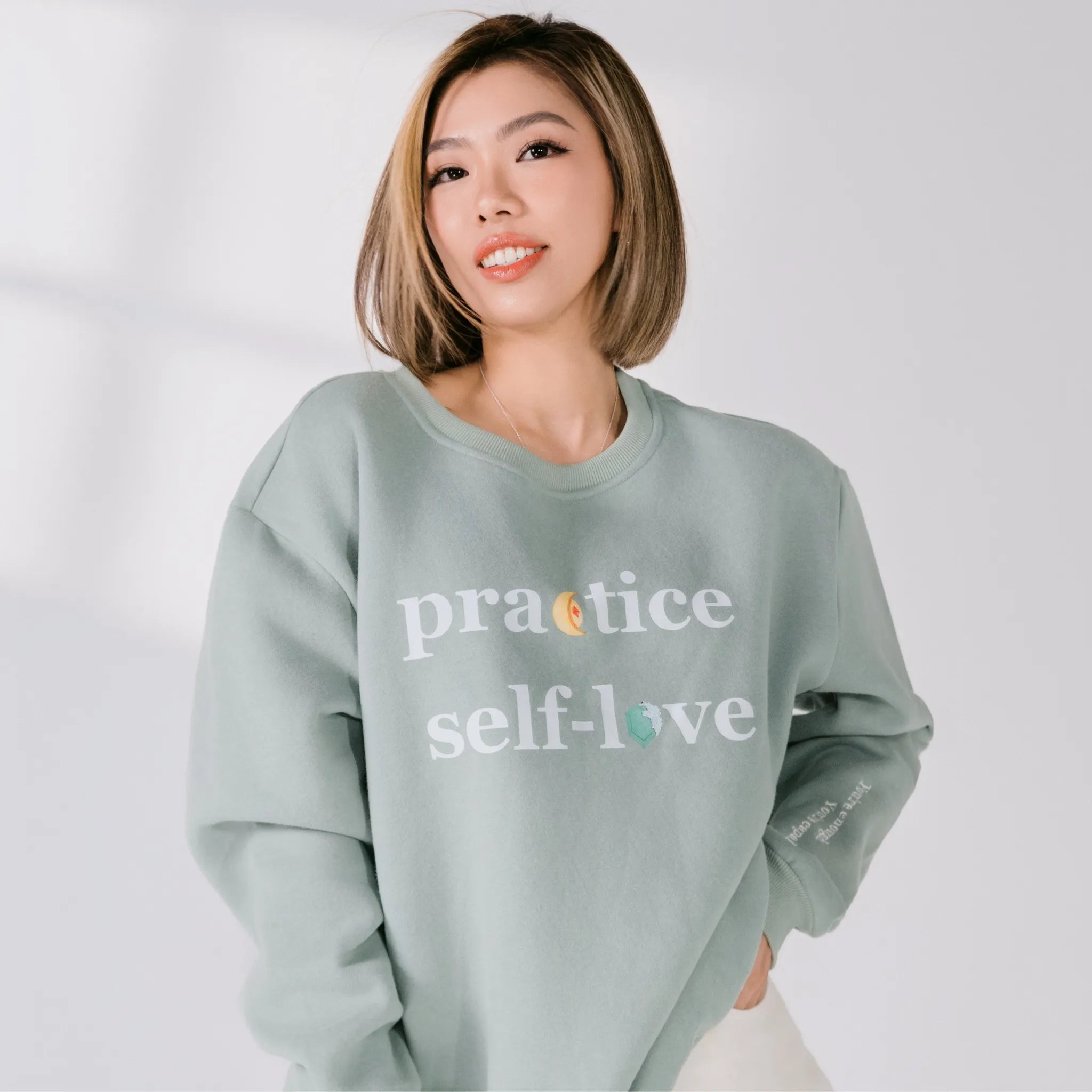 Person wearing a light green sweatshirt with 'practice self-love' text on a plain background