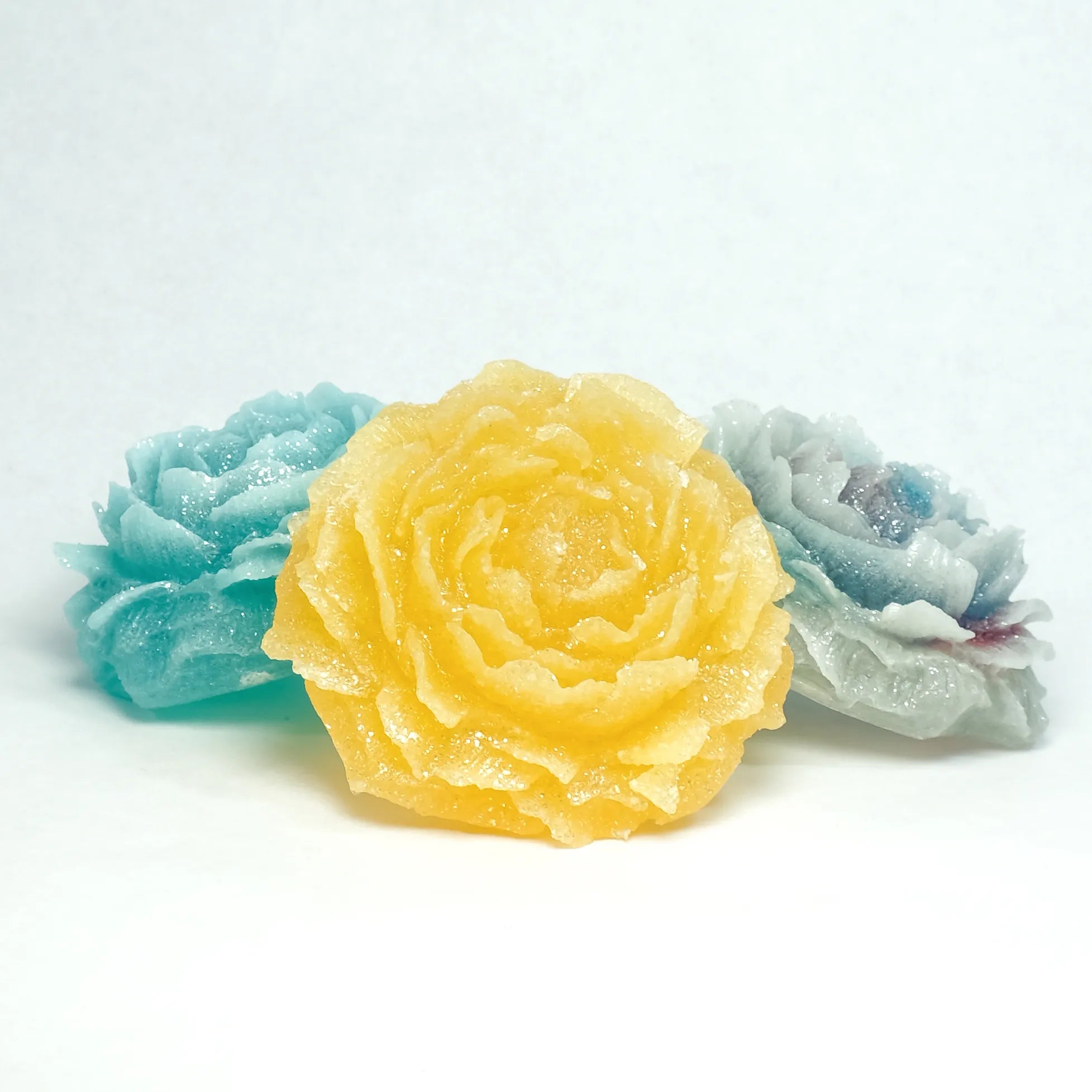 Three floral-shaped soaps in yellow, blue, and white on a light gray background