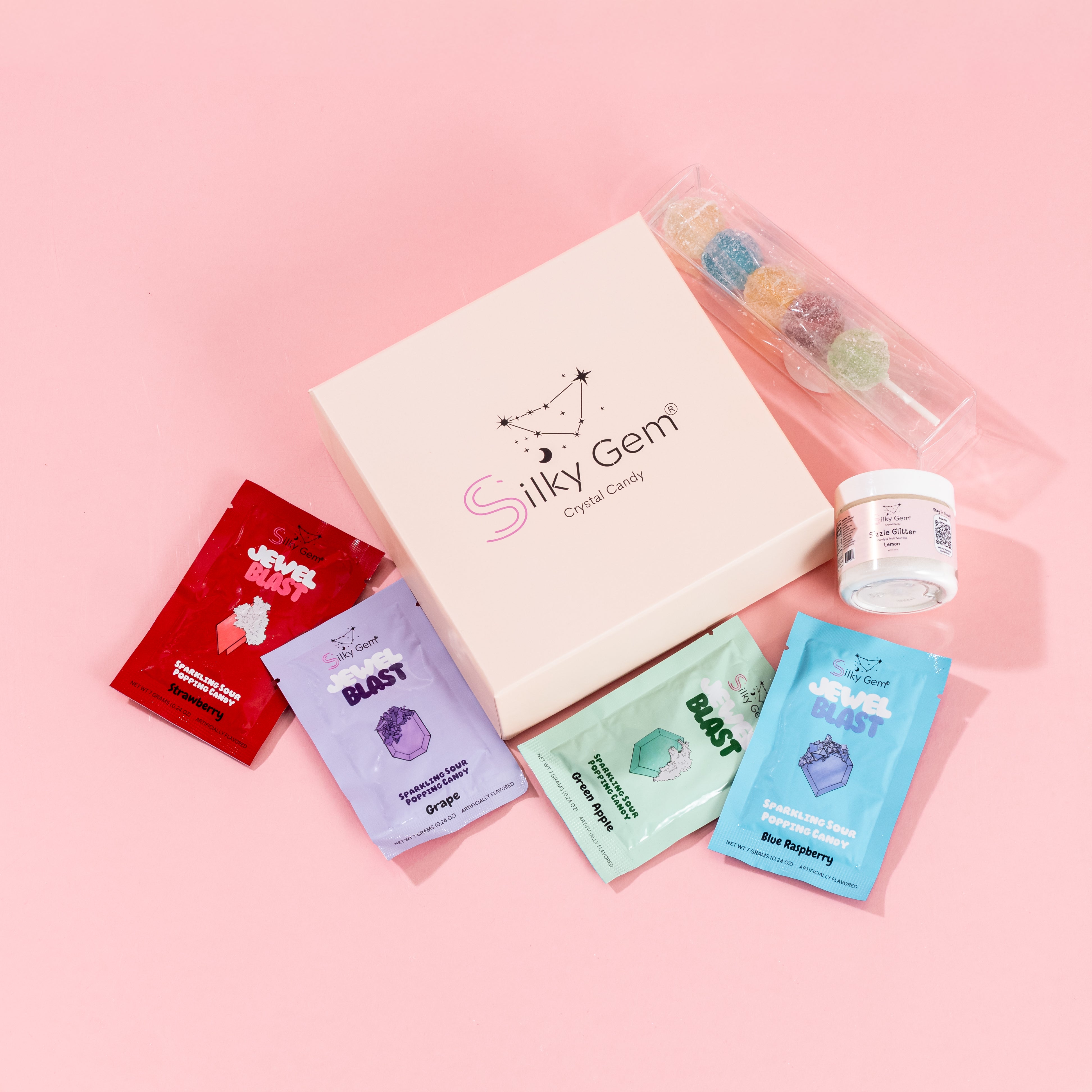 Silky Gem® Starter Kit bundle including the branded Treasure Box collection inside its gift box, a Sizzle Pop skewer, lemon-flavored Sizzle Glitter, and four pouches of Jewel Blast sparkling sour popping candy, all arranged on a pink background.