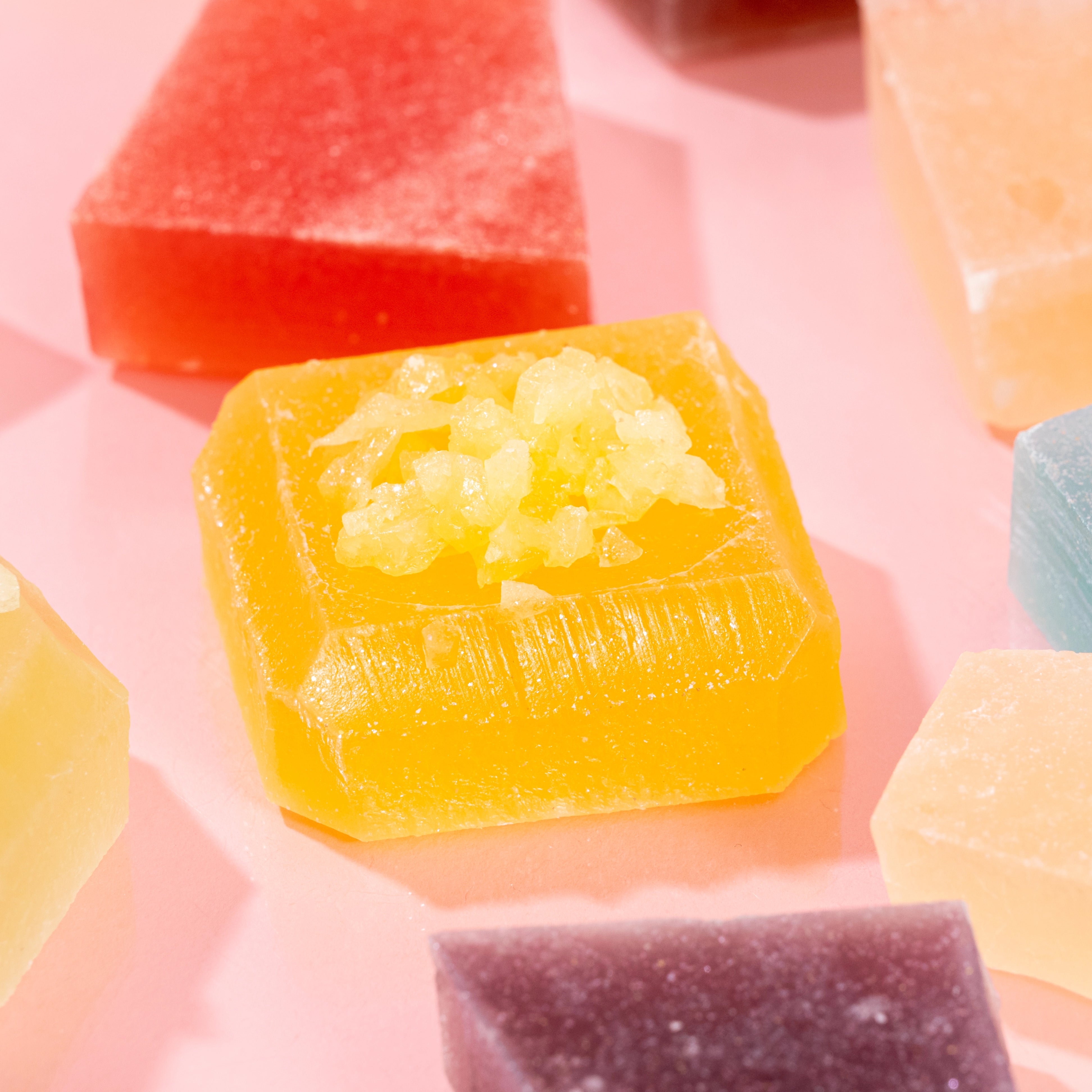 Close-up of vibrant, crystal-cut candy pieces from the Treasure Box collection, with some pieces cut in half to reveal the signature firm, jelly-like center, displayed on a pink background.