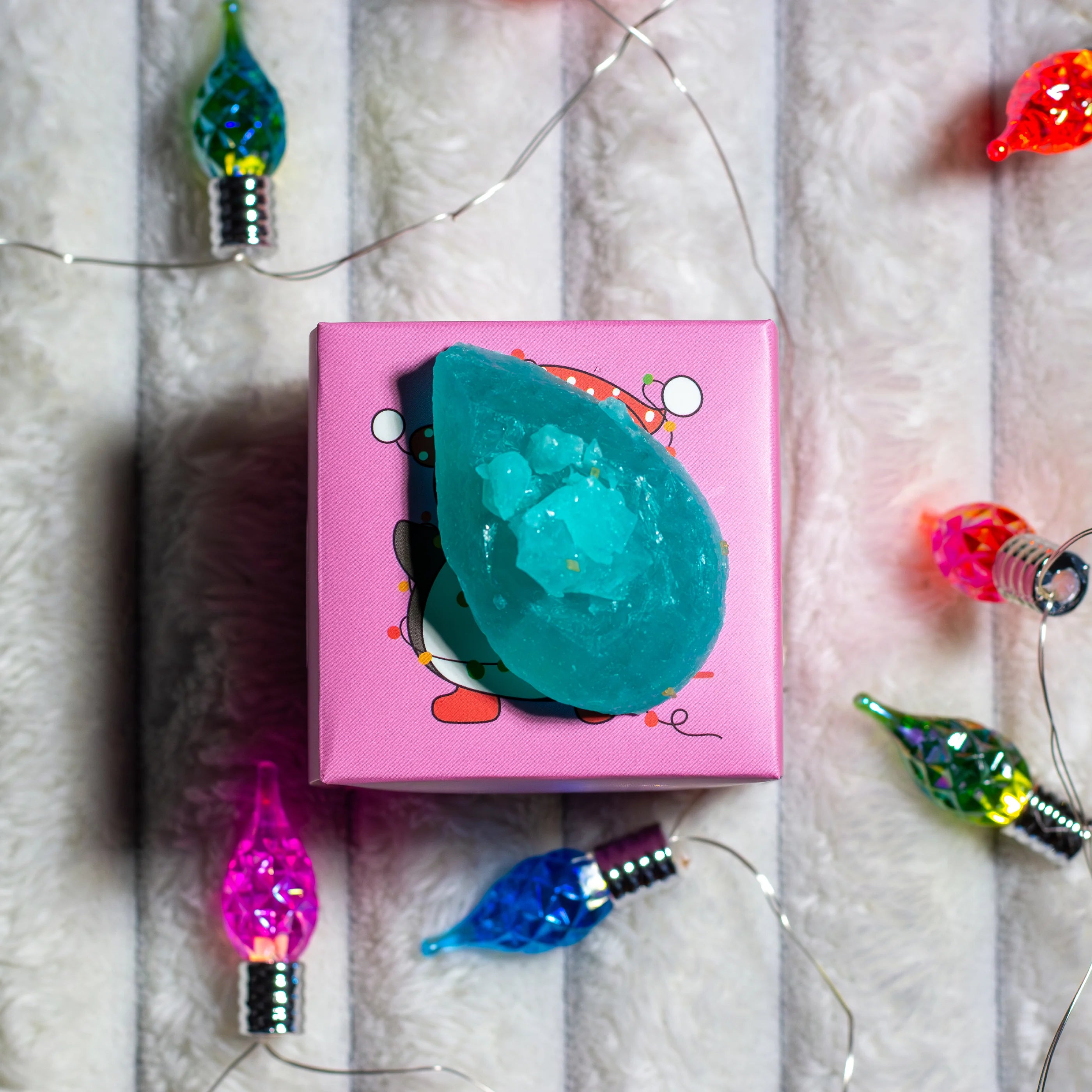 Pink box with a teal bath bomb on a textured surface with colorful string lights.