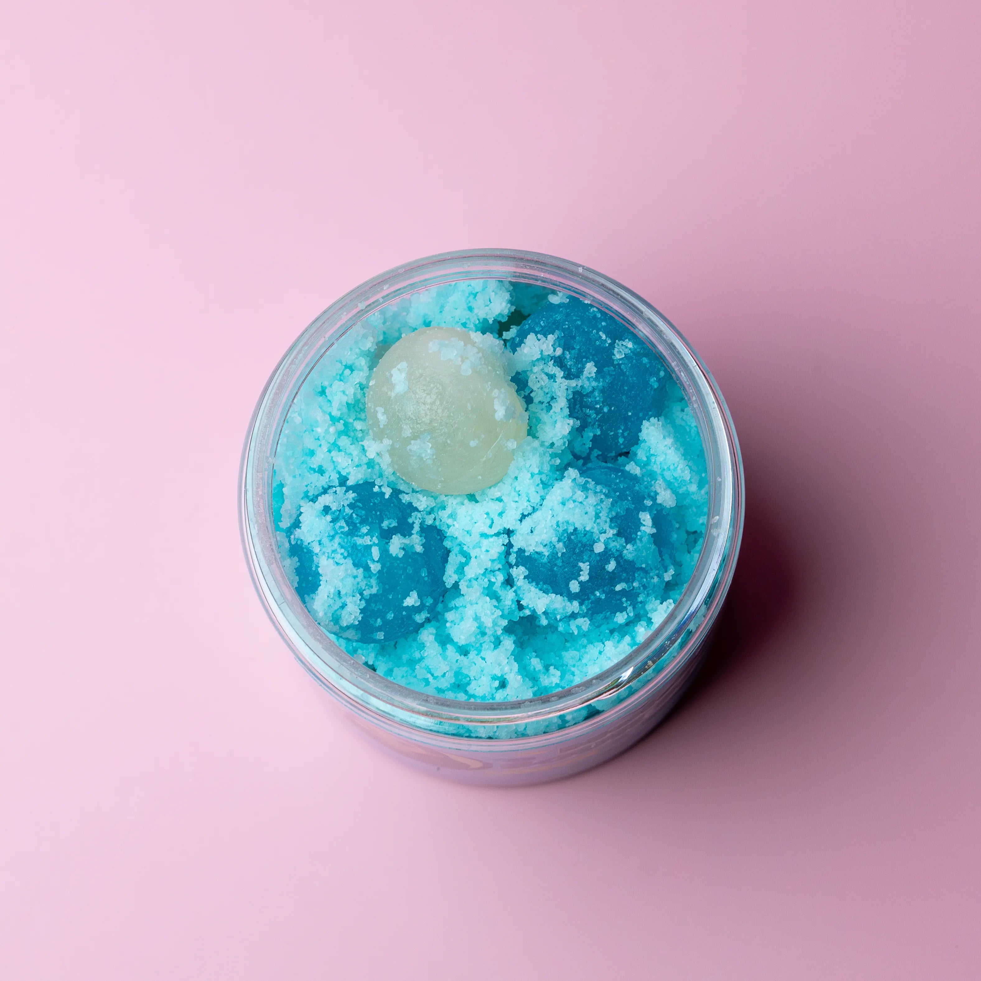 Jar of blue bath salt with a green bath bomb on a pink background