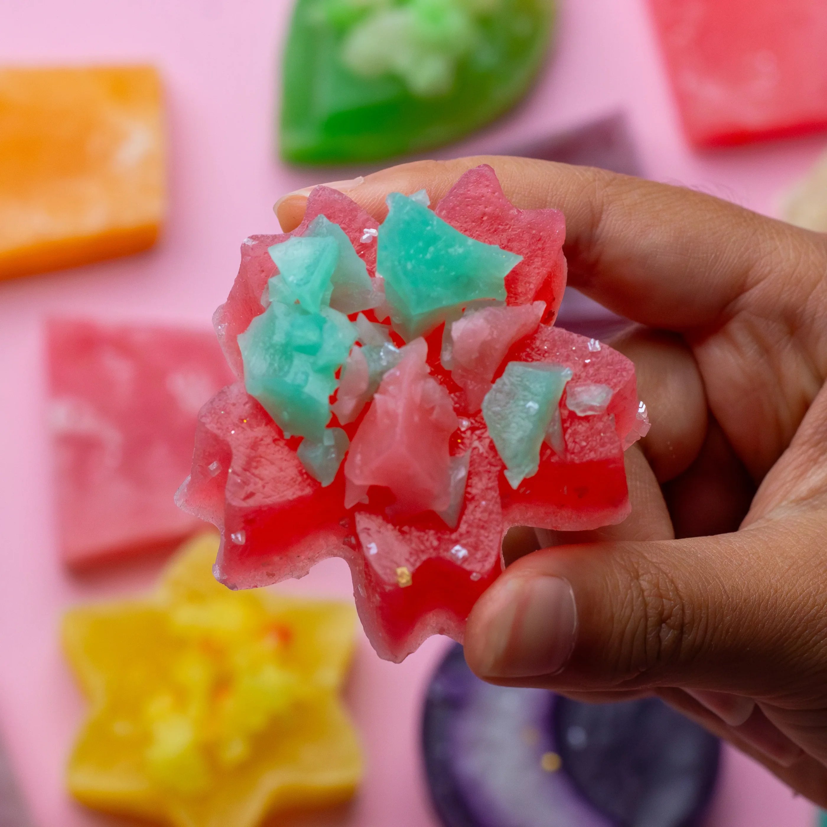 Colorful gummy candy shaped like a flower held by a hand against a pink background with more gummy candies.