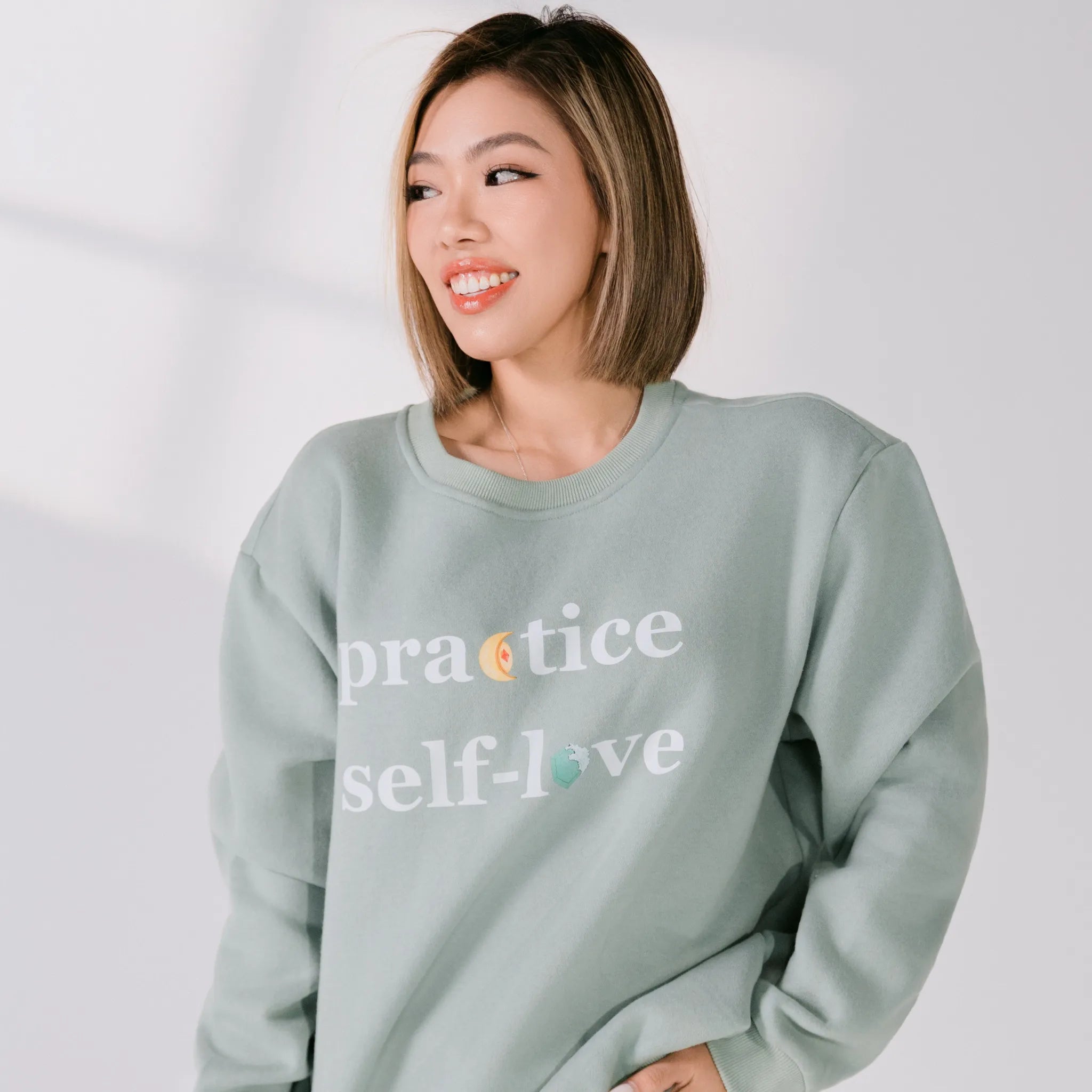 Person wearing a light green sweatshirt with 'practice self-love' text on a plain background
