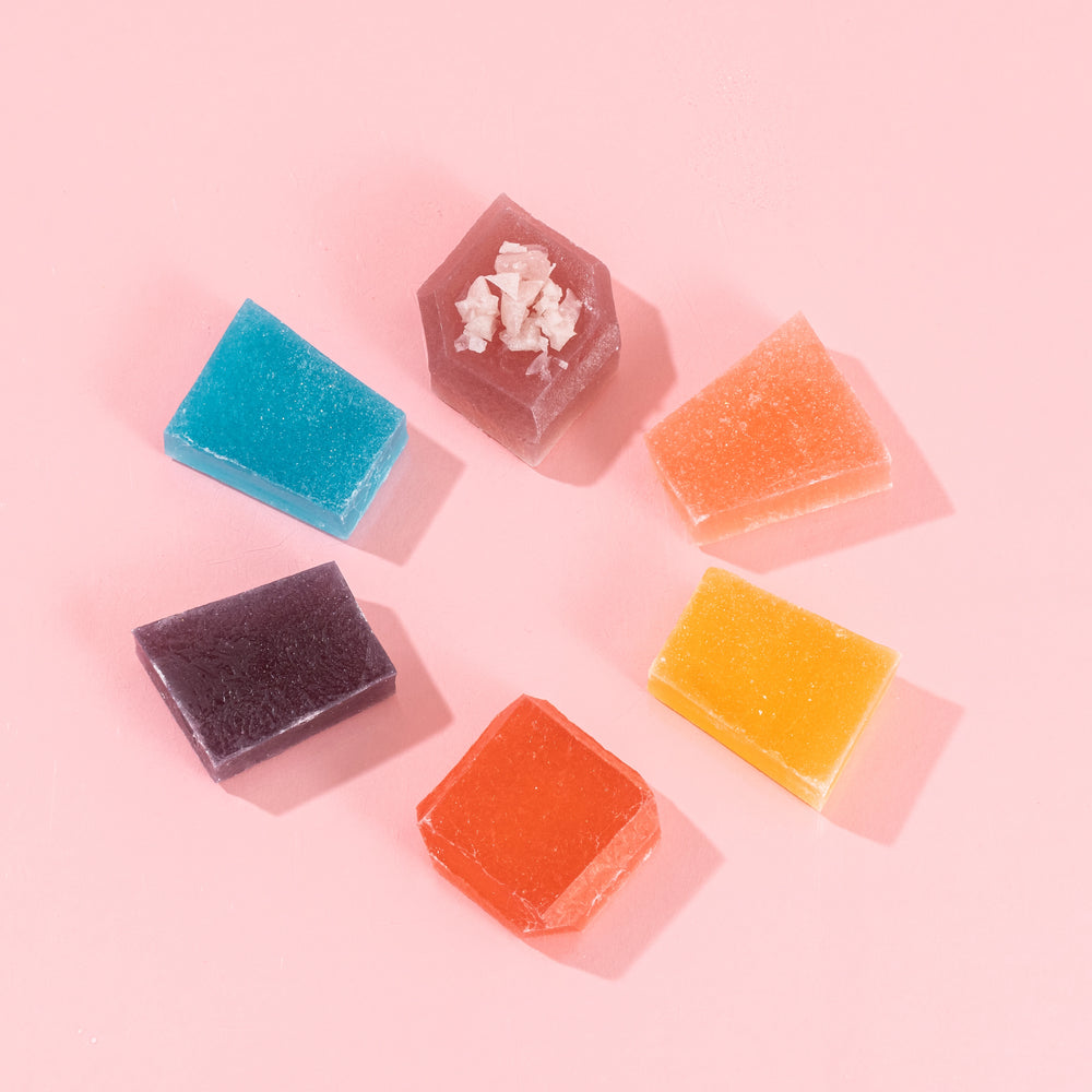 Assorted Silky Gem® crystal candies in various shapes and colors arranged in a circular pattern on a pink background, with one piece featuring a light dusting of Sizzle Glitter.