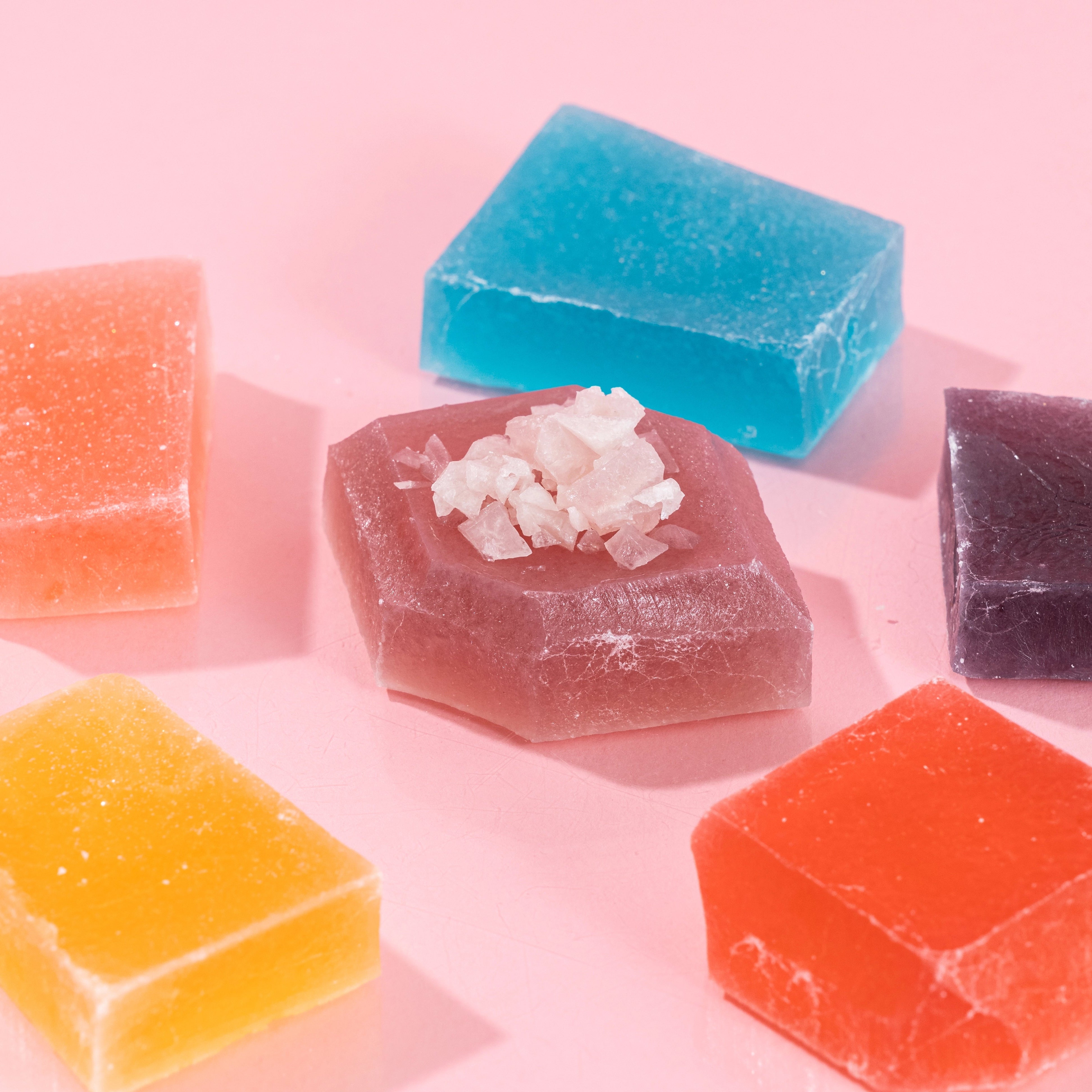 Five colorful Silky Gem® crystal candies in various geometric shapes arranged on a pink background.