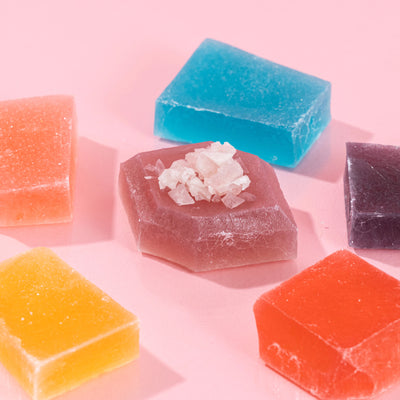 Five colorful Silky Gem® crystal candies in various geometric shapes arranged on a pink background.