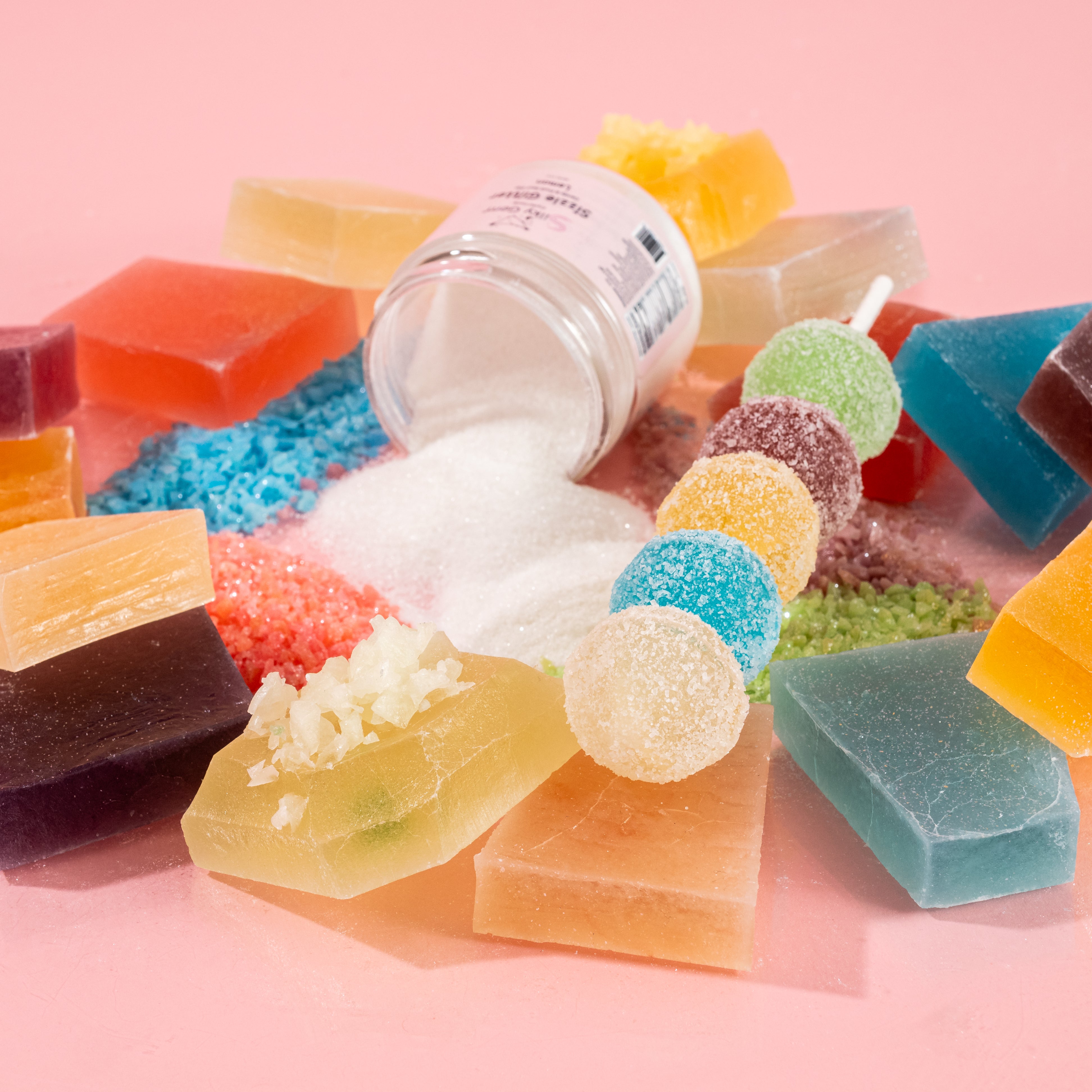 Assorted Silky Gem crystal candies, including pieces with signature cuts and a jar of Sizzle Glitter, arranged artfully on a pink background.