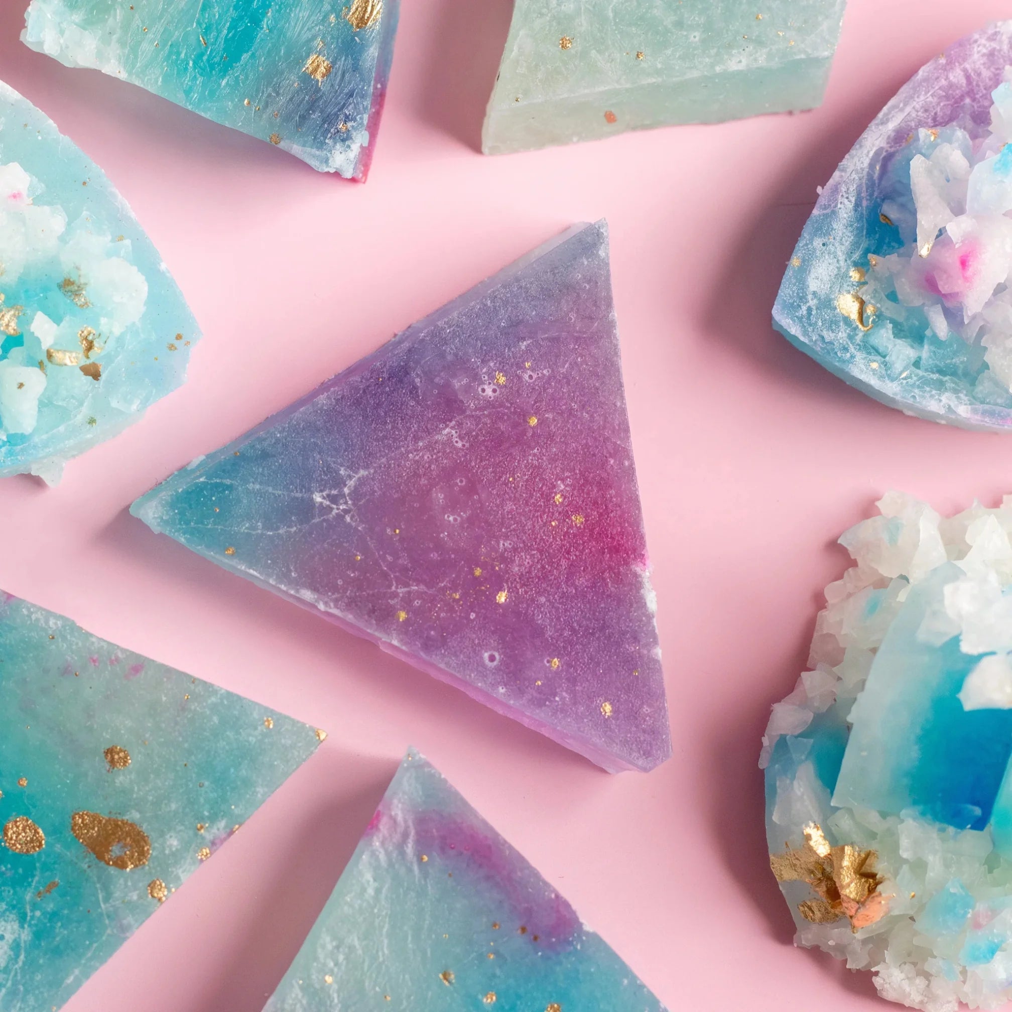 Assorted geometric-shaped lychee aurora flavored crystal candies in various shades of purple, blue, and pink, with gold glitter, displayed on a pink background.